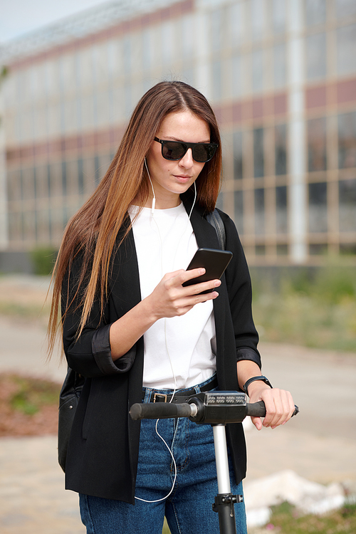 Contemporary young woman in earphones and casualwear scrolling through playlist in smartphone while moving to work on scooter