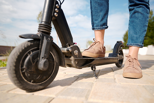 Legs of young woman in beige shoes and blue jeans standing on electric scooter on road against in urban environment and cloudy sky