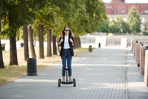 Happy young casual female in sunglasses moving on electric scooter along road between riverside and row of green trees in the morning