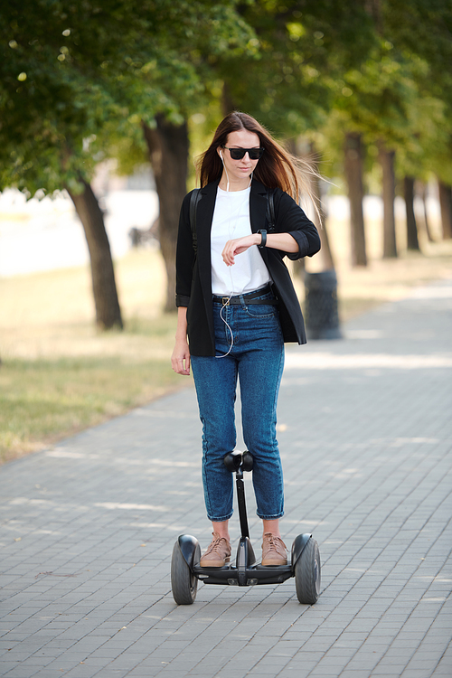 Young contemporary elegant businesswoman looking at wristwatch while riding on gyroscope along road against green trees growing in row