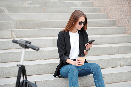 Young casual woman with long hair sitting on staircase in urban environment and scrolling in smartphone while listening to music