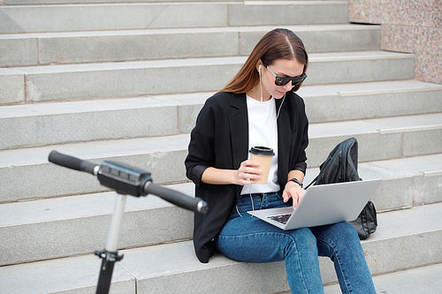 Contemporary casual girl with glass of coffee looking at laptop display while surfing in the net on staircase in urban environment