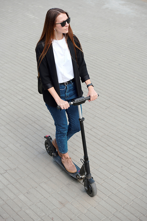 Young stylish woman in sunglasses, jeans, cardigan and pullover standing on electric scooter while moving down asphalt road in the city