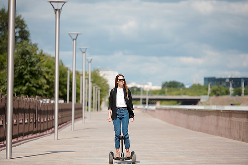 Young long-haired woman in sunglasses and smart casualwear standing on gyroscope while moving along road against cloudy sky and riverside
