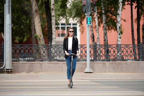 Young elegant woman in sunglasses and casualwear standing on electric scooter and moving down crosswalk while green lights are on