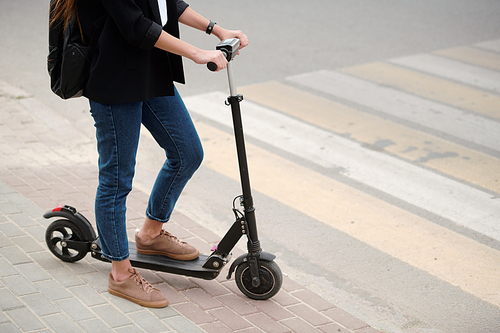 Low section of young female in blue jeans and black jacket keeping one leg on electric scooter while standing by road with crosswalk