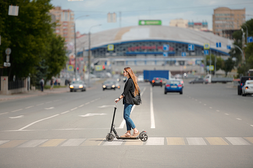 Side view of young female in casualwear standing on electric scooter and moving down crosswalk against trade center and other buildings
