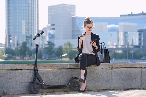 Young relaxed businesswoman with smartphone and coffee sitting by riverside with electric scooter near by against modern architecture
