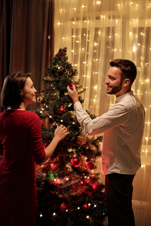 Happy young brunette woman in smart red dress looking at her husband putting decorations on Christmas tree during discussion at home