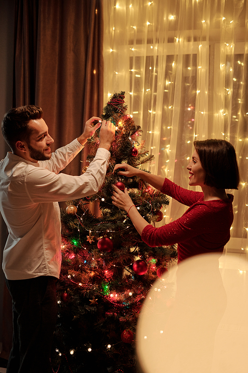 Happy young brunette woman in smart red dress and her bearded husband putting decorations on Christmas tree during conversation
