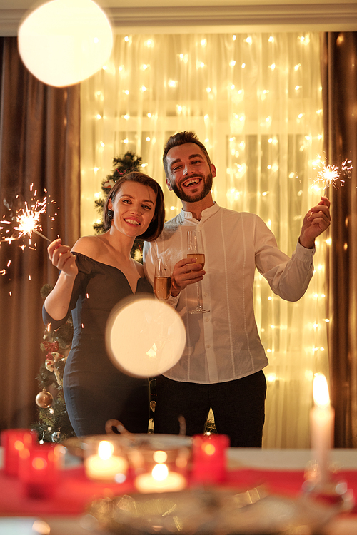 Young cheerful elegant couple with bengal lights in hands standing against decorated xmas tree in living-room during home party and looking at you