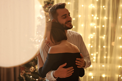 Happy young bearded man in white shirt embracing his elegant wife in black evening dress during dance at Christmas home party against xmas tree
