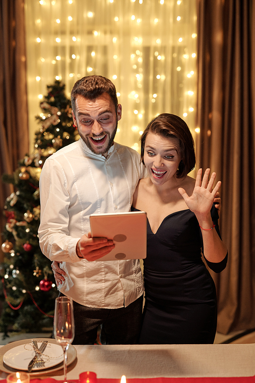 Young cheerful elegant couple looking at smartphone screen while communicating to someone in video chat and wife waving hand