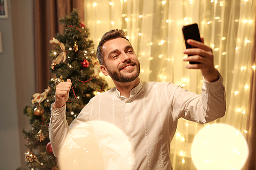 Smiling handsome young man in white shirt pointing at Christmas tree while taking selfie in living room
