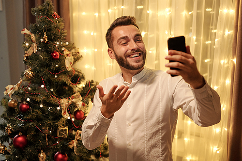 Young cheerful bearded man in white shirt looking at smartphone screen while making selfie or communicating to someone against Christmas tree