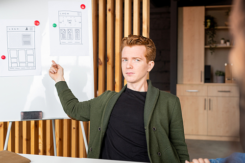 Young confident developer of mobile software pointing at papers with sketches on whiteboard while explaining details to colleagues