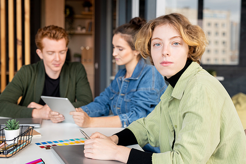 Young blond serious female mobile app developer looking at you while sitting by workplace against her two colleagues using touchpad