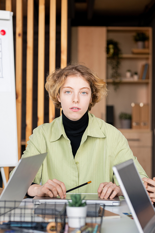 Young blond serious female mobile app developer looking at you while sitting by workplace between two laptops used by her colleagues