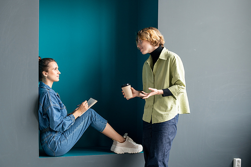 Two contemporary young businesswomen in casualwear discussing work while one of them sitting against wall and her friend having drink