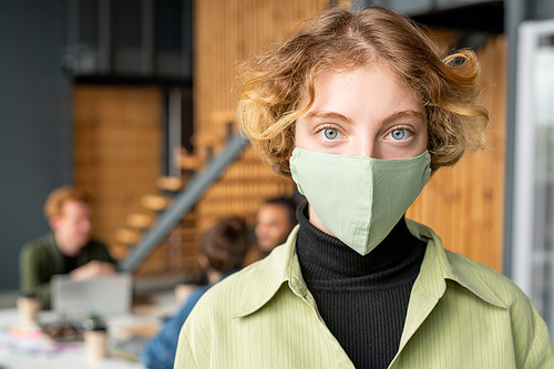 Young blond female office worker in casualwear and protective mask looking at you while standing in front of camera against colleagues