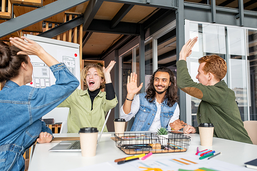 Four young excited intercultural designers giving each other high five while sitting by desk in office and celebrating success of their new project