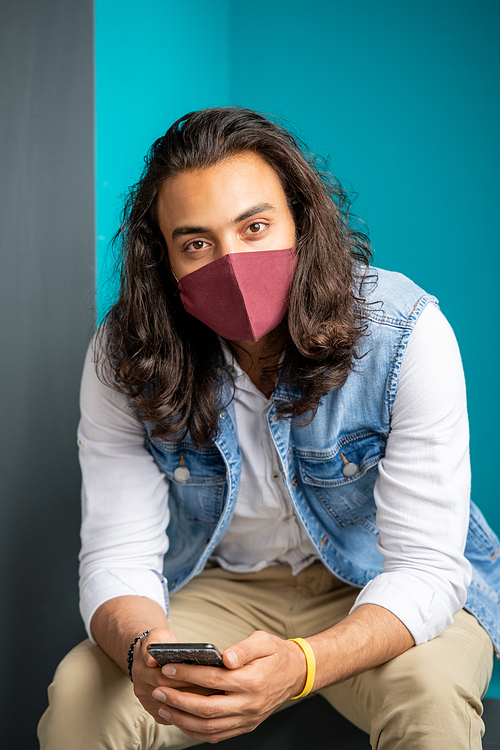 Young mixed-race businessman in casualwear and protective mask looking at you while sitting against blue wall in office and using smartphone