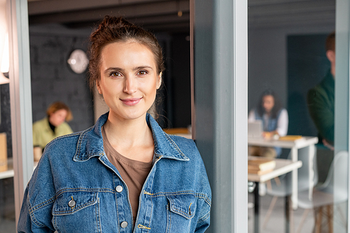 Young successful female office manager in denim shirt looking at you while standing against open door and her colleagues working