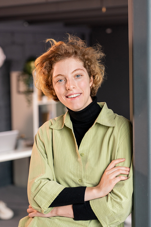 Young smiling blond female office worker in casualwear standing by open door in front of camera and looking at you against workplace