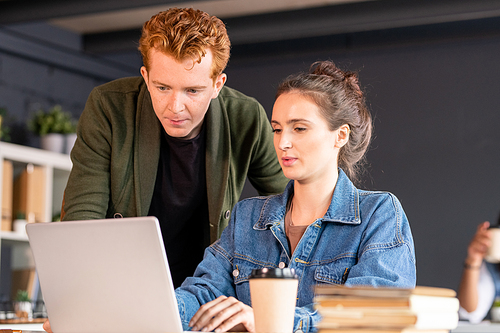 Young confident brunette female designer and her serious male colleague looking at laptop display while woman making presentation