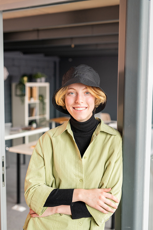 Young cheerful blond businesswoman in casualwear looking at you with toothy smile while standing by open door with office on background