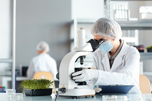 Portrait of female scientist looking in microscope while studying plant samples in biotechnology lab, copy space