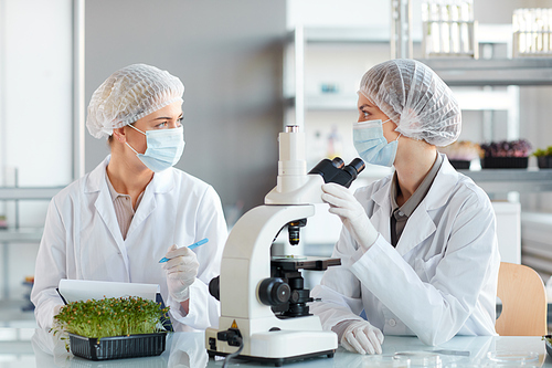 Portrait of two young female scientists looking in microscope while studying plant samples in biotechnology lab, copy space