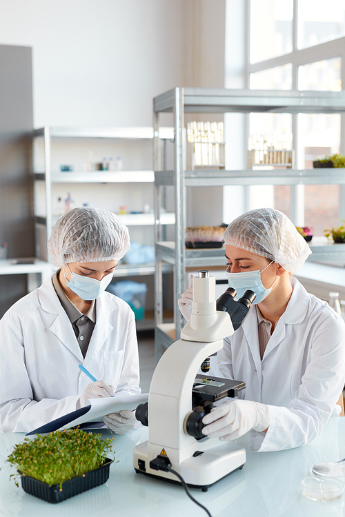 Vertical portrait of two young female scientists looking in microscope while studying plant samples in biotechnology lab