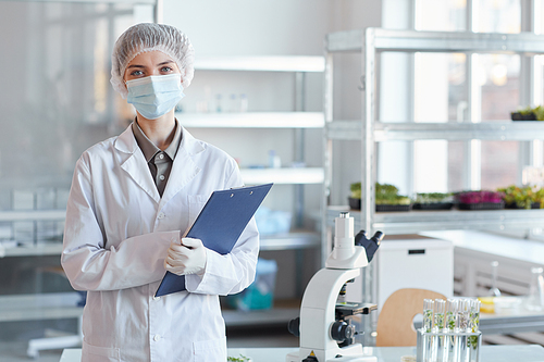 Waist up portrait of young female scientist wearing face mask and looking at camera while standing in medial laboratory and holding clipboard, copy space