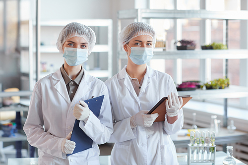 Waist up portrait of two female scientists wearing face mask and holding clipboards while standing in medical laboratory