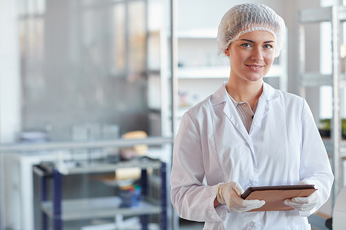 Waist up portrait of young female scientist holding digital tablet and smiling at camera while standing in medical laboratory, copy space