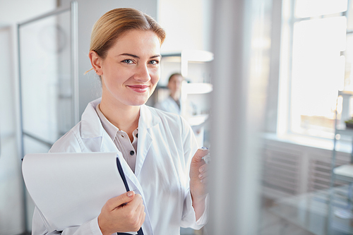 Waist up portrait of young female scientist smiling at camera and holding clipboard while standing in medical laboratory, copy space