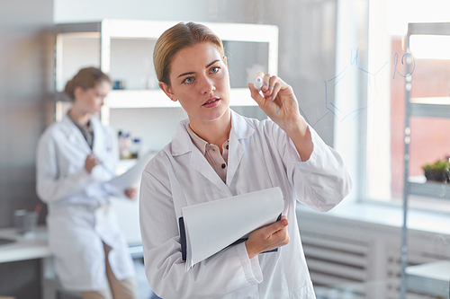 Waist up portrait of young female scientist writing on glass wall and holding clipboard while standing in medical laboratory, copy space