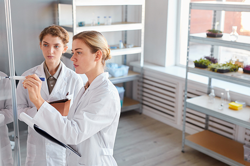 Side view portrait of two female scientists writing on glass wall and holding clipboard while doing research in medical laboratory, copy space