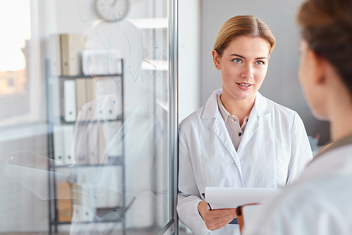 Waist up portrait of two female scientists talking and holding clipboard while doing research in medical laboratory, copy space