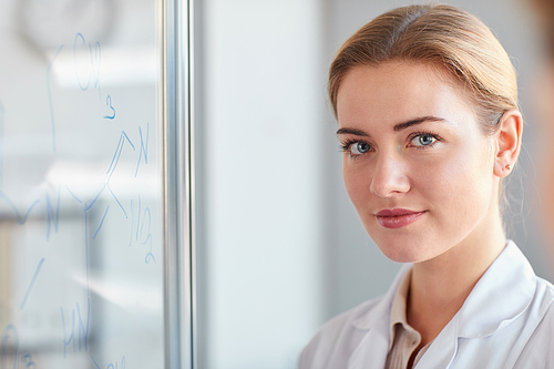 Close up portrait of blonde female scientist looking at camera while standing by glass wall with writings in medical laboratory, copy space