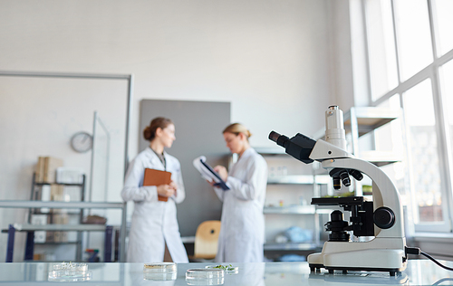 Portrait of two female scientists discussing research while standing in medical laboratory, focus on microscope in foreground, copy space