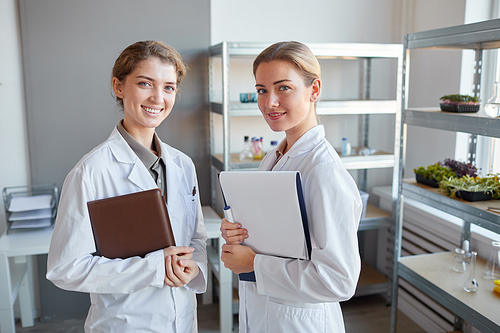 Waist up portrait of two young female scientists looking at camera and smiling while standing n medical laboratory, copy space