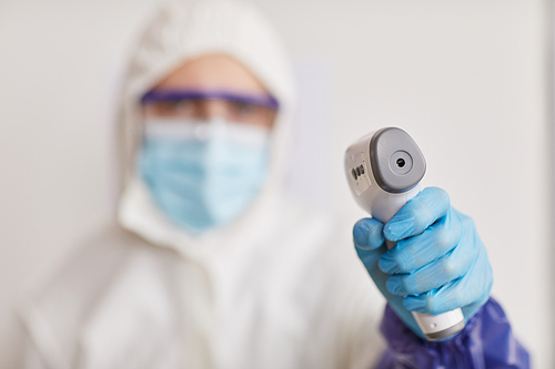 Close up of medical worker wearing full protective gear and pointing contsctless thermometer at camera while taking temperature, focus on foreground, copy space