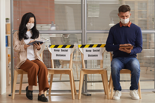 Full length portrait of young people wearing masks while waiting in line in office with Keep Social Distance signs, copy space