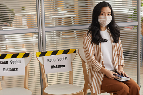 Portrait of young Asian woman wearing mask and looking at camera while waiting in line with Keep Social Distance signs, copy space