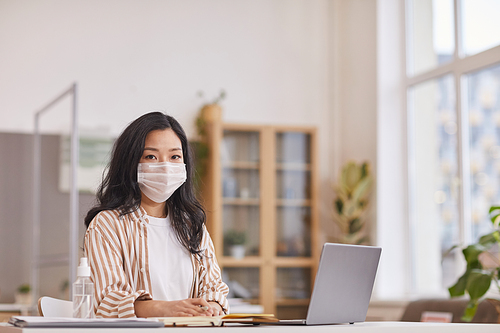 Portrait of young Asian woman wearing mask and looking at camera while sitting at desk in office with bottle of sanitizer in foreground, copy space