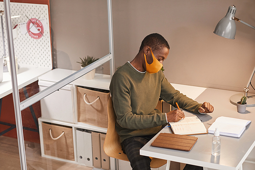 High angle portrait of young African-American man wearing mask and writing in planner while working at desk in office cubicle, copy space
