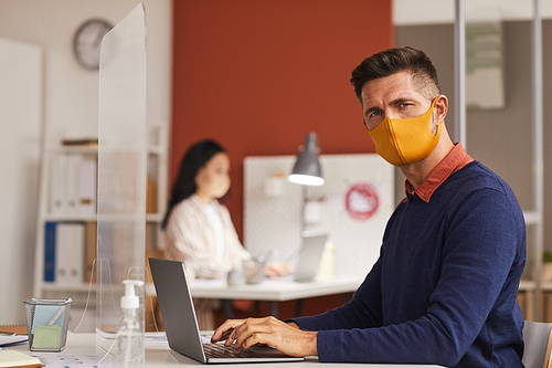 Portrait of modern mature man wearing mask and looking at camera while using laptop at desk in office, copy space