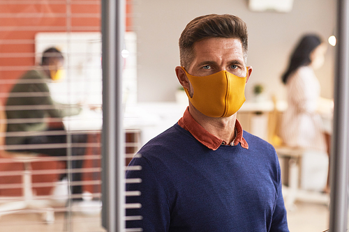 Waist up portrait of handsome mature man wearing mask and looking at camera while standing in office interior, copy space
